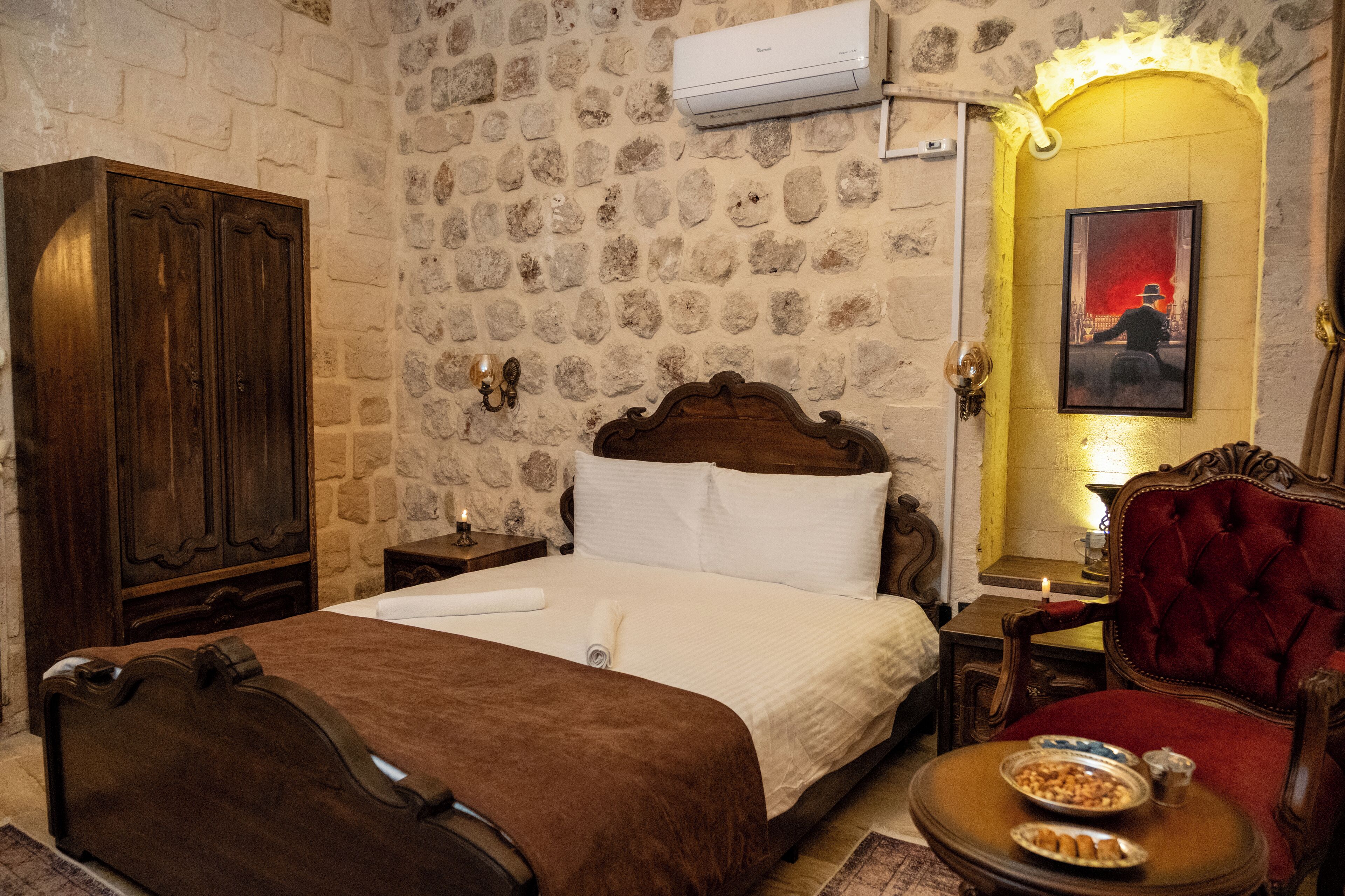 Une chambre d’hôtel avec un mur en pierre, un lit en bois, un fauteuil rouge et une petite table avec des en-cas.