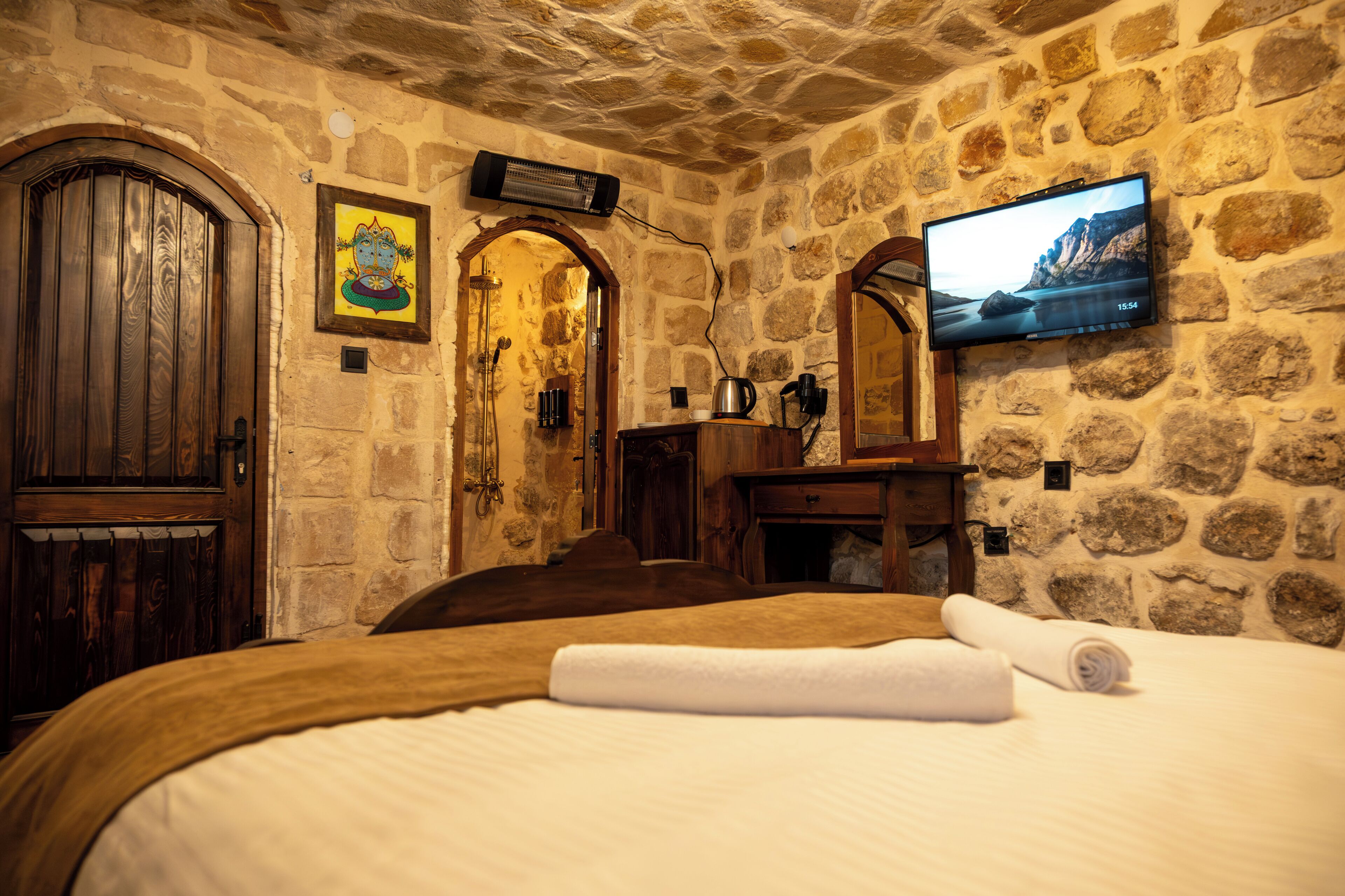 A stone-walled room with a bed, a wooden door, a painting, a television, and a small table with a kettle.