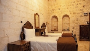 A room with two beds, a wooden nightstand, a mirror, and a stone wall. - Marinut Konağı (Mardin)
