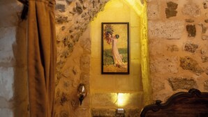 A stone-walled room with a framed painting, a wooden bed, and a wall-mounted light fixture. - Marinut Konağı (Mardin)