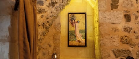 A stone-walled room with a framed painting, a wooden bed, and a wall-mounted light fixture.