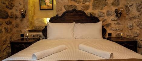 A bed with white linens and rolled towels, set against a stone wall with a painting above.