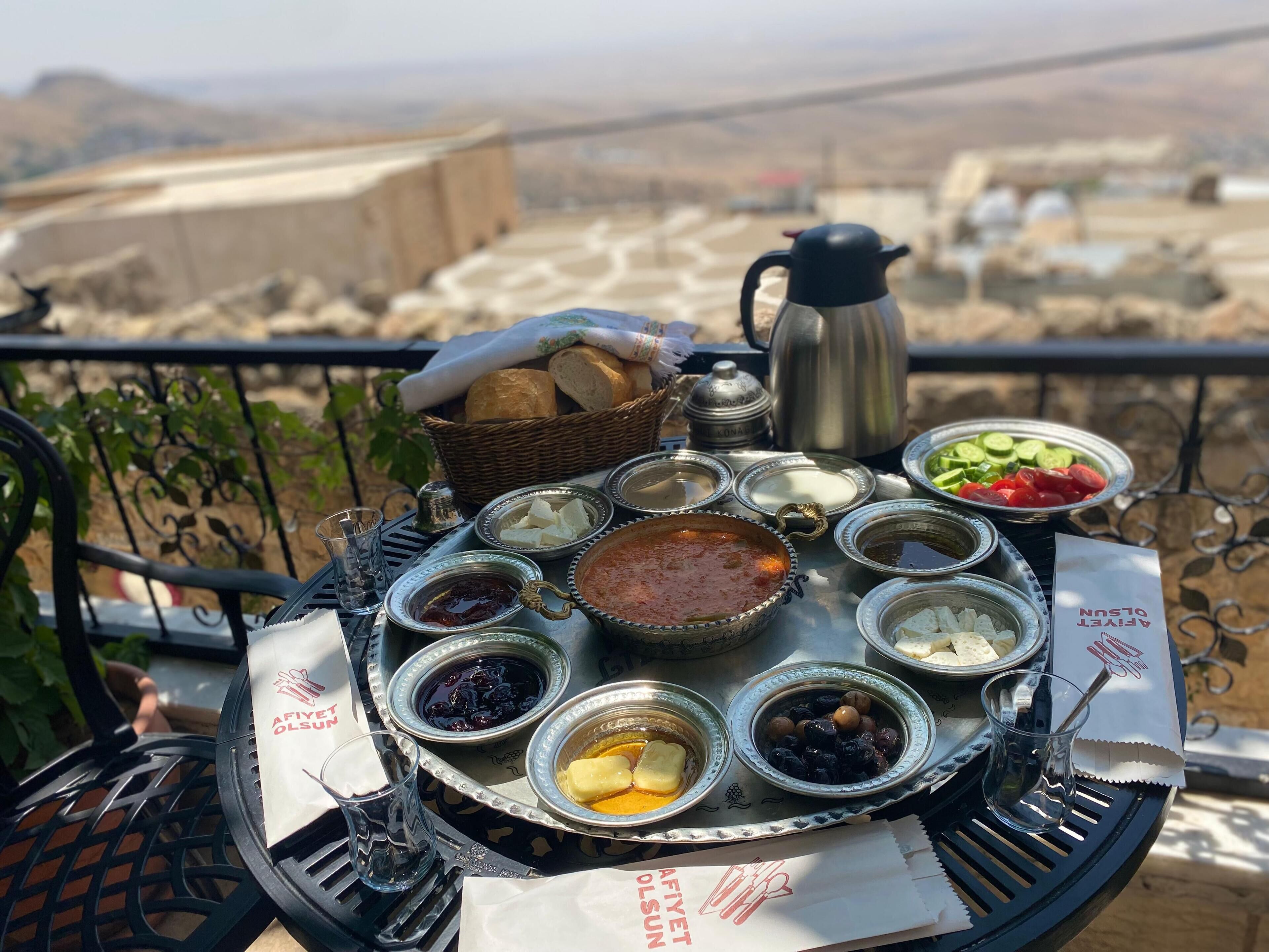 A table with various food items and a kettle, set on a balcony overlooking a scenic view.
