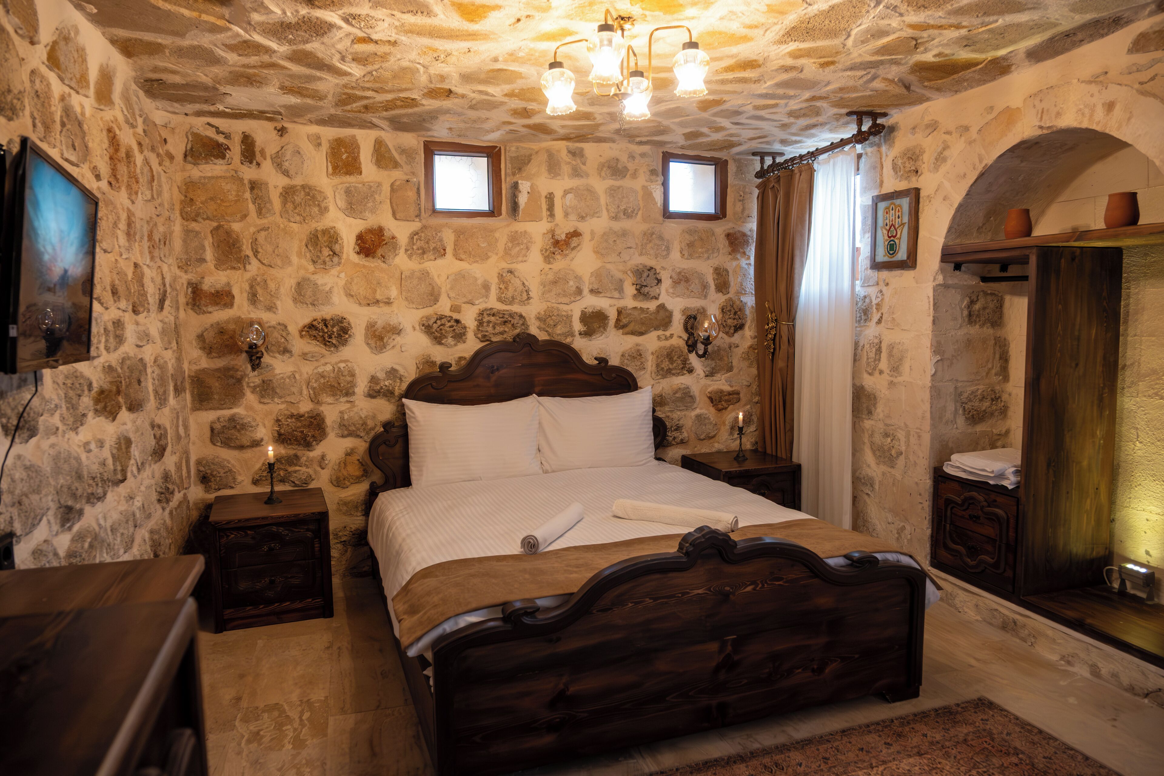 Un dormitorio con paredes de piedra, una cama de madera, dos mesitas de noche, un pequeño ventanal y una repisa con una vela.