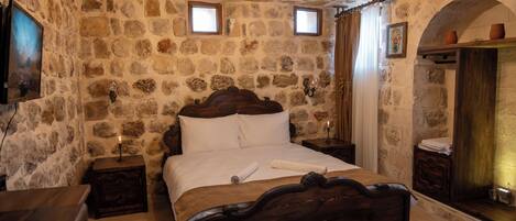 A stone-walled bedroom with a wooden bed, two bedside tables, a nightstand, a small window, and a wall-mounted shelf with a candle.
