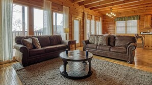 A cozy log cabin interior with two leather sofas, a round coffee table, and large windows with curtains.
