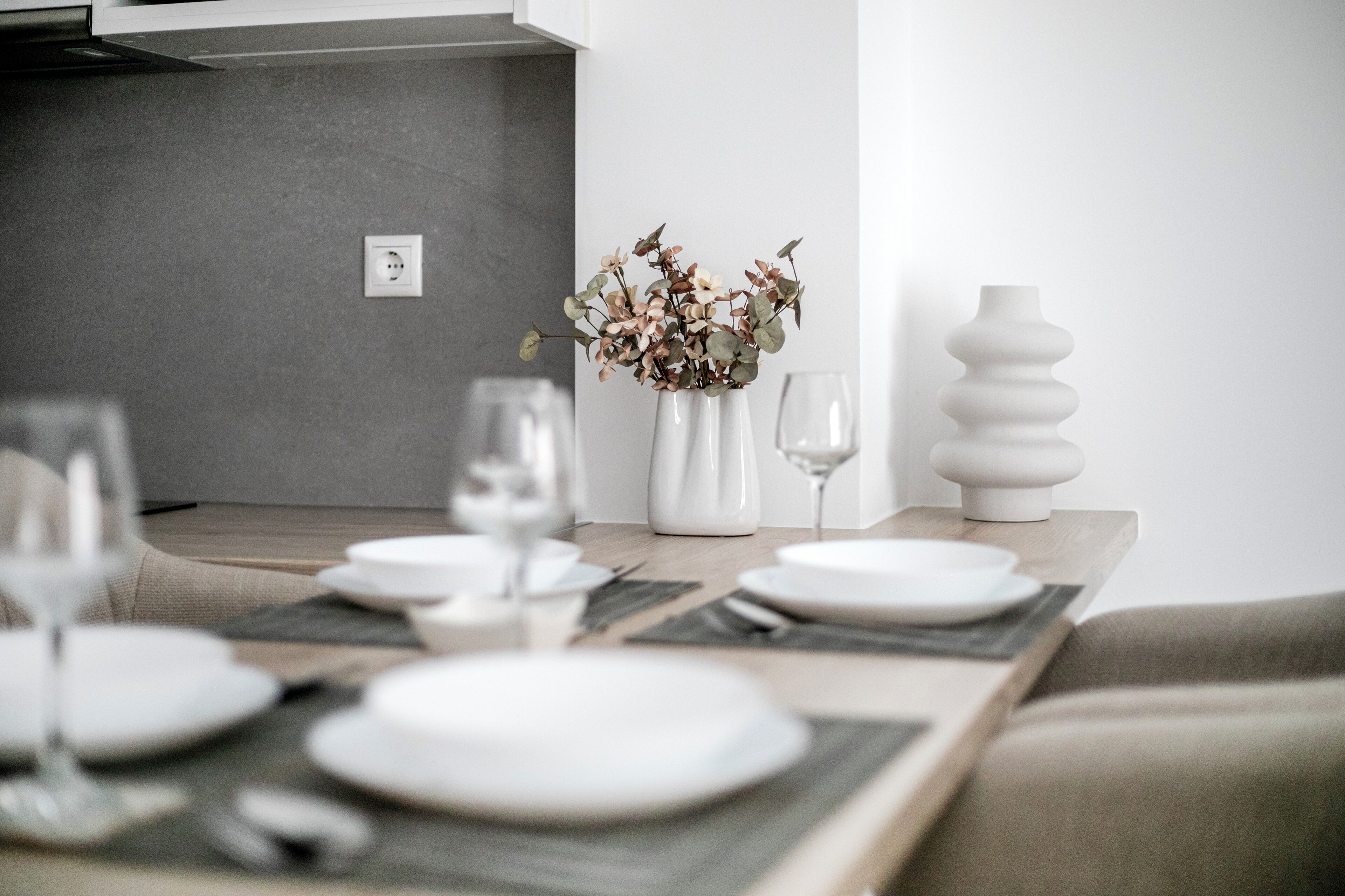 Une table à manger dressée avec de l’assiette blanche, des verres et un vase contenant des fleurs séchées.