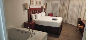 A hotel room with a large bed, a wooden headboard, a bedside table with a lamp, a red chair, a wooden desk, and a large bathtub.