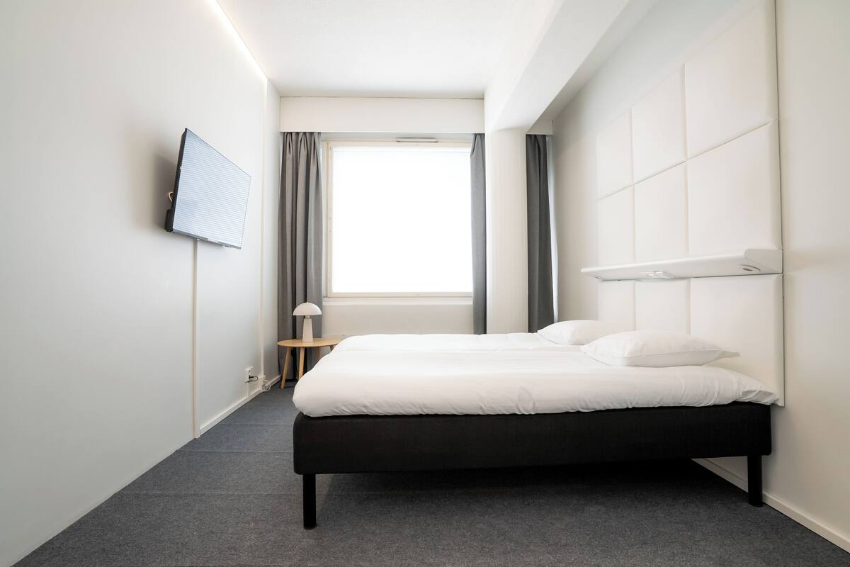 Une chambre d’hôtel moderne équipée d’un lit, d’un téléviseur à écran plat fixé au mur, d’une fenêtre avec des rideaux et d’une petite table avec une lampe.