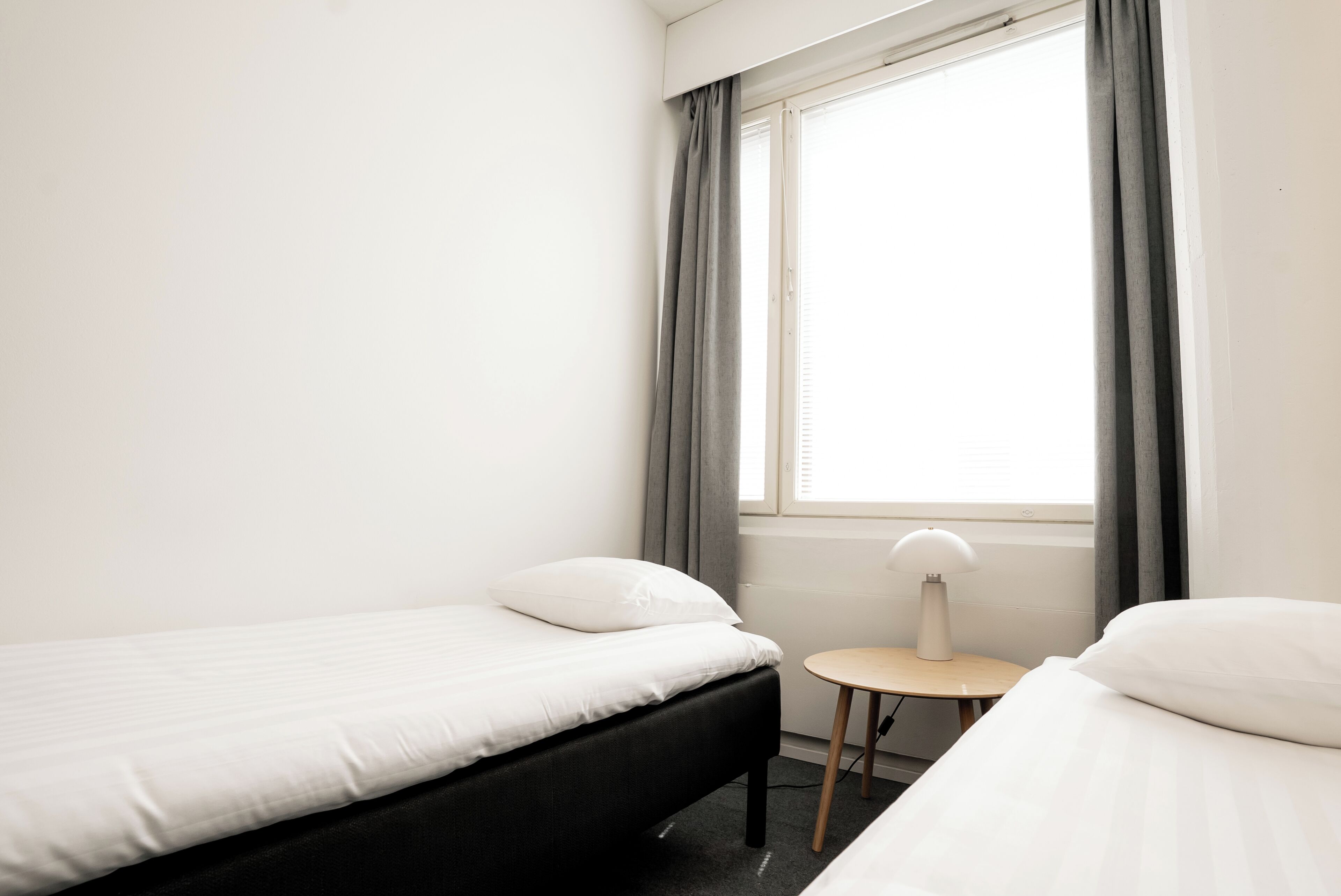 A small, clean room with two single beds, a small table with a lamp, and a window with curtains.