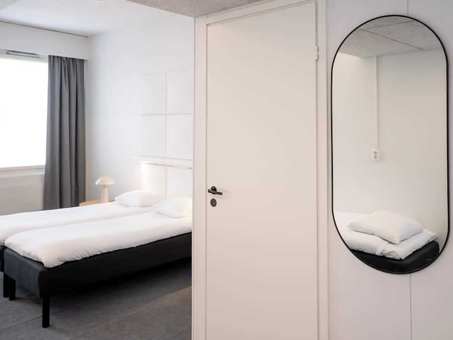 Une chambre à coucher avec un lit double, une table de chevet avec une lampe, un grand miroir et une fenêtre avec des rideaux.