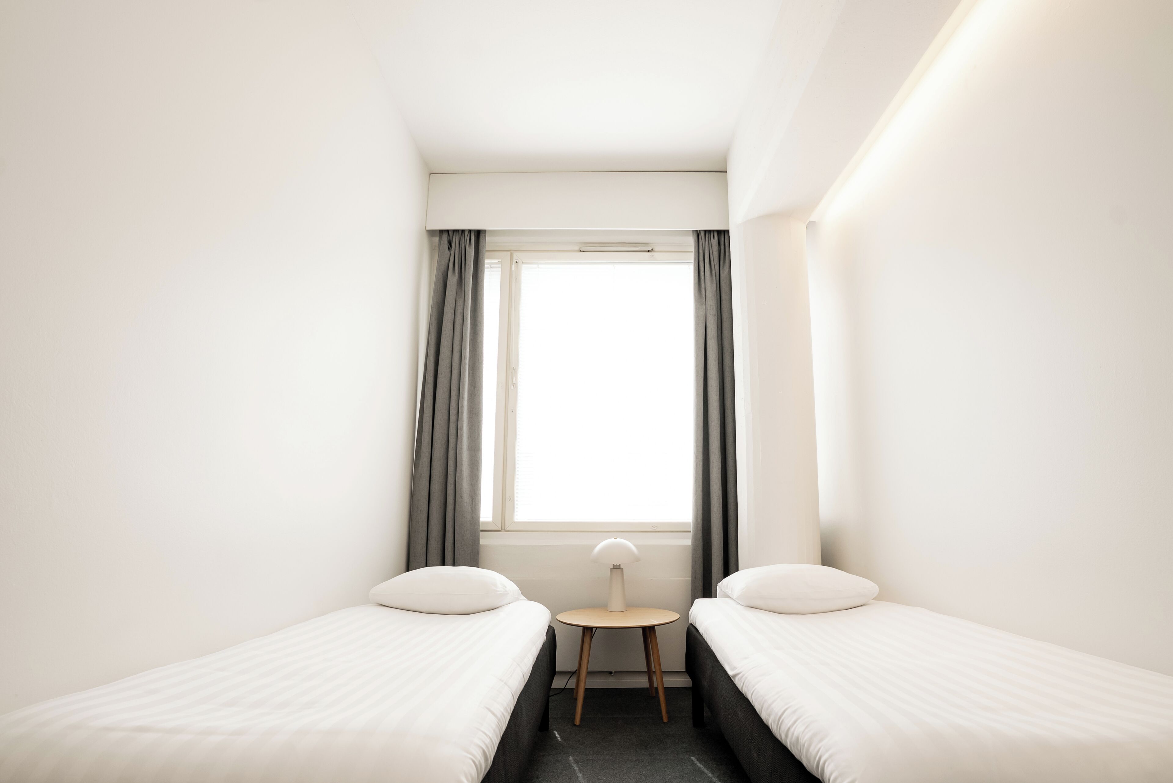 A small, modern hotel room with two single beds, a small table, and a window with curtains.