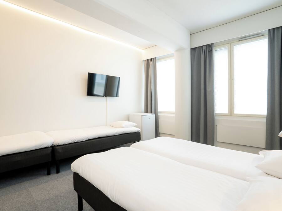 A hotel room with two beds, a flat-screen TV mounted on the wall, a bedside table with a lamp, and a window with curtains.