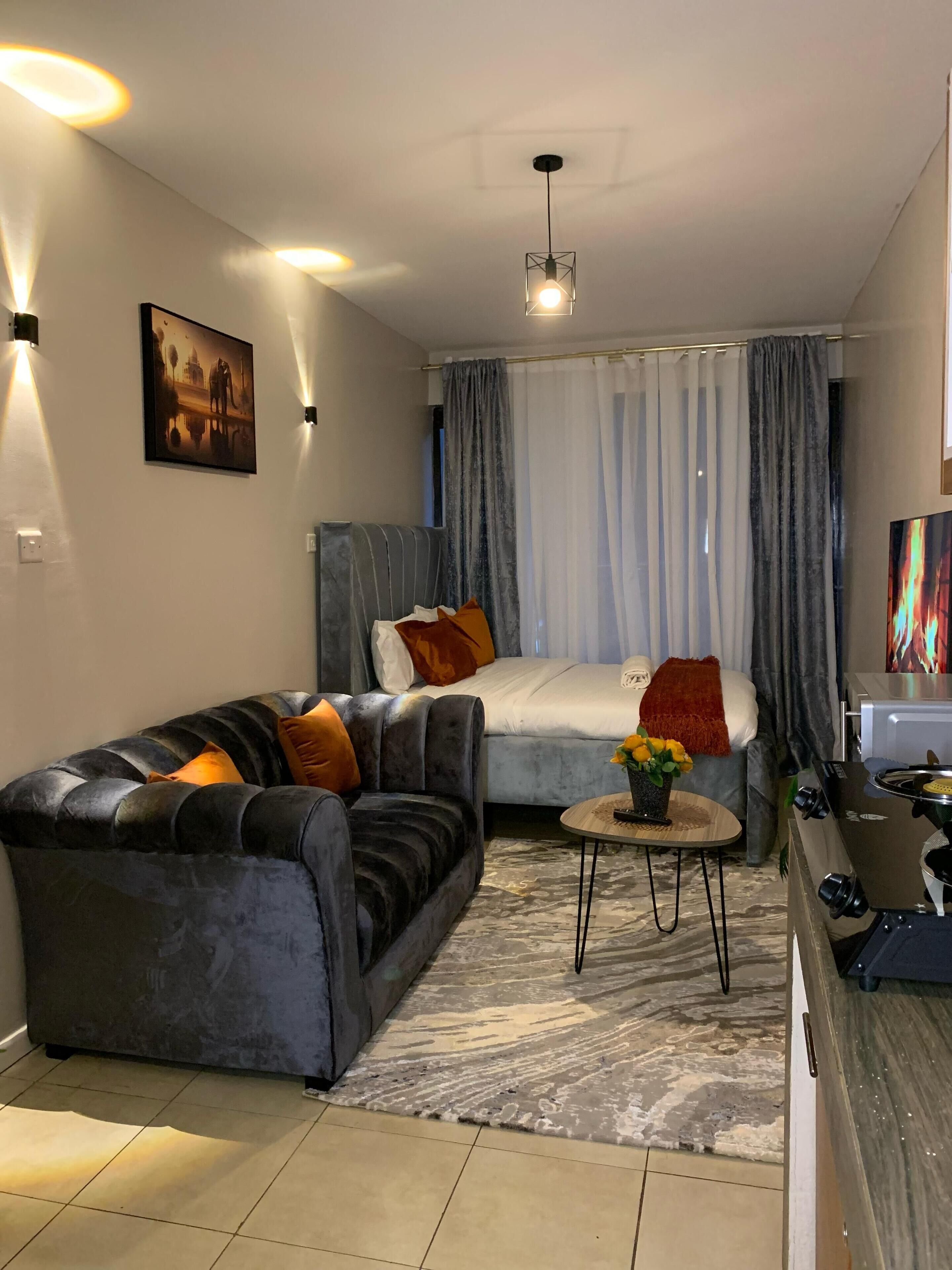 A modern living room with a grey sofa, a bed with grey curtains, a small table with a vase of flowers, and a kitchen area with a stove.