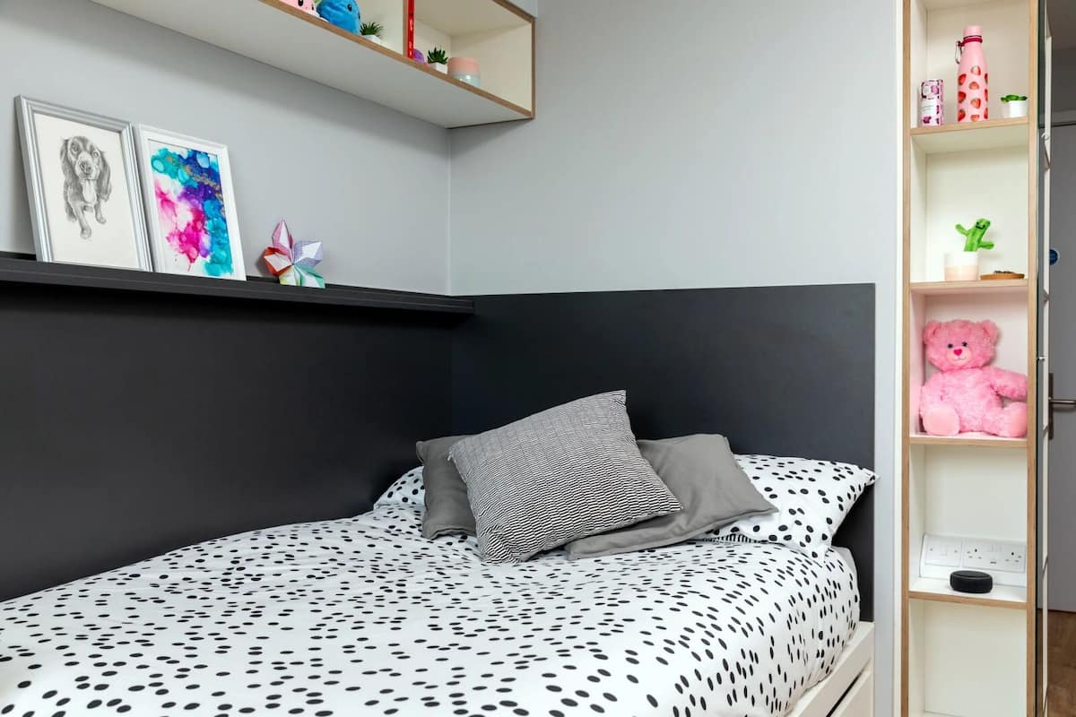A bedroom with a bed, pillows, a shelf with items, and a wall with artwork.