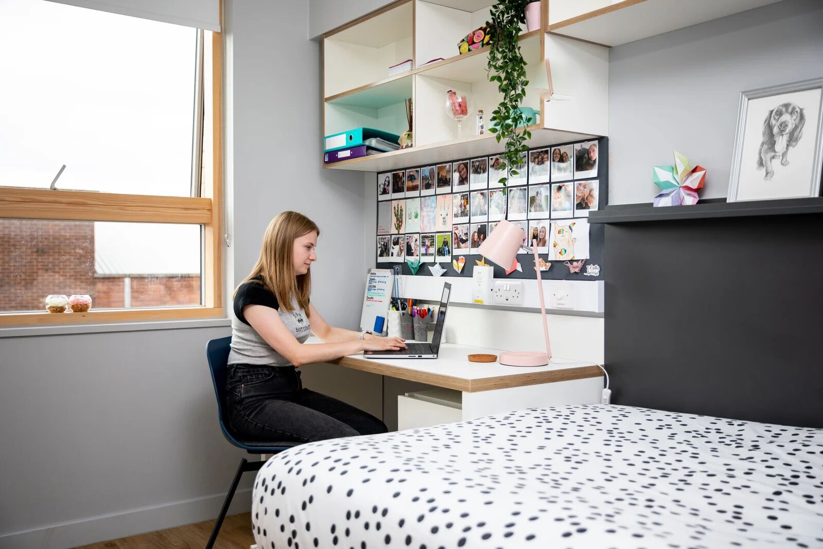 Una habitación con escritorio, laptop, cama con cubrecamas de lunares, ventana y estante con fotos y una planta.
