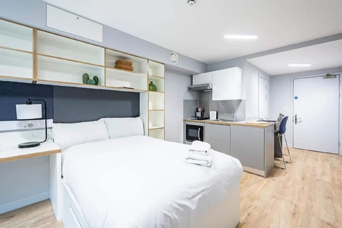 A modern hotel room with a large bed, a desk with a computer, a kitchenette, and a bathroom.