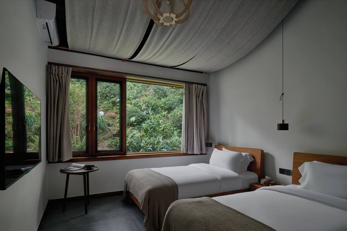 A hotel room with two beds, a small table, a window with curtains, and a ceiling fan.