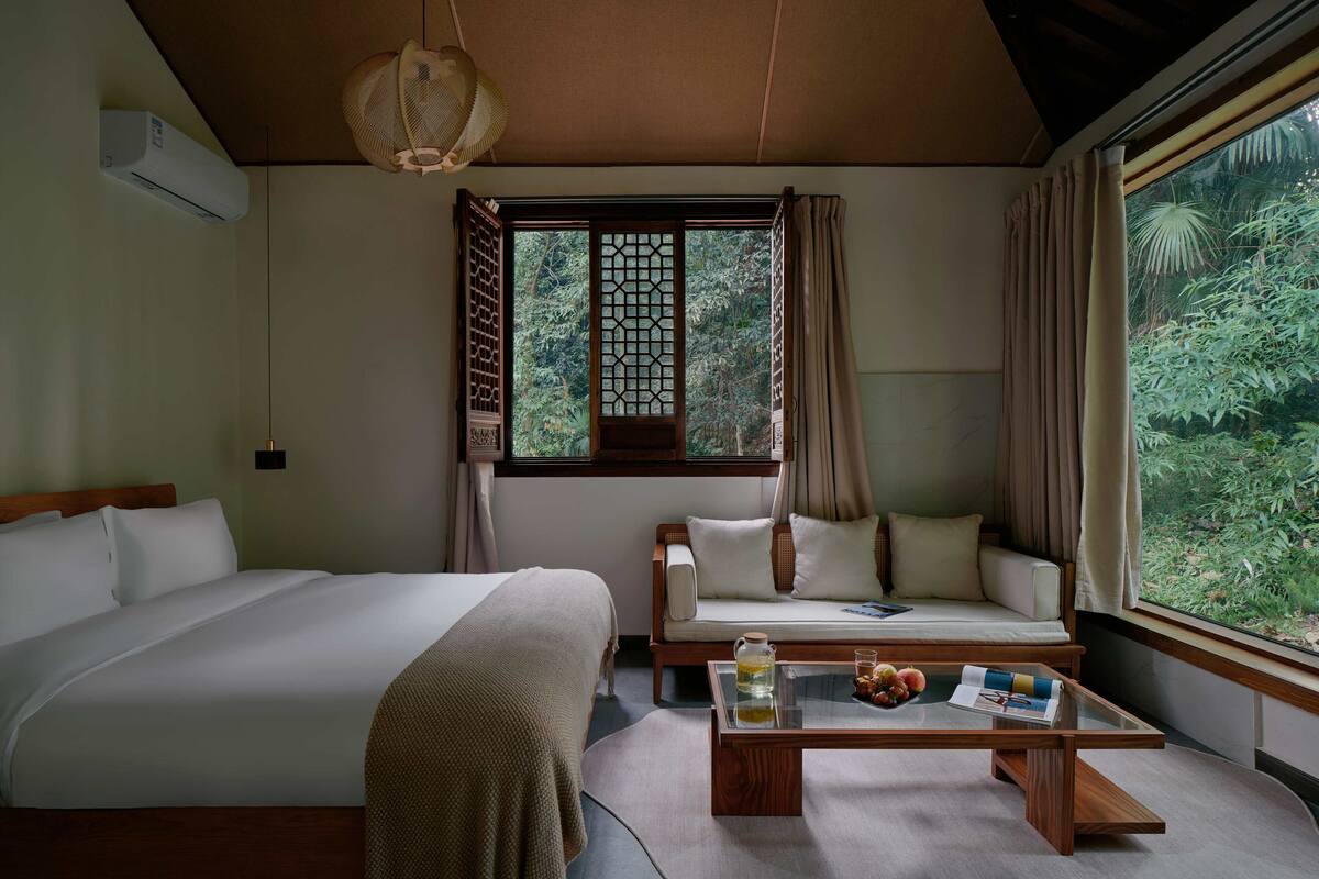 A bedroom with a large bed, a sofa, a glass coffee table, and a window with a view of greenery.