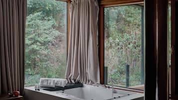 A bathroom with a bathtub, a window with curtains, and a view of greenery outside.