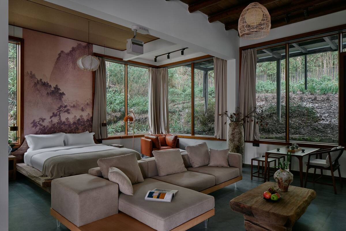 A modern bedroom with a large bed, a sofa, a dining table, and a view of the forest through large windows.