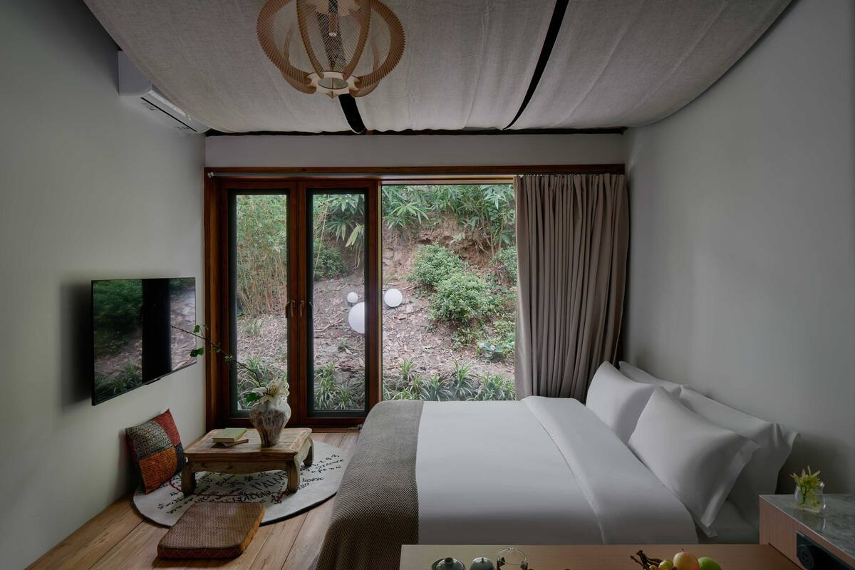 A bedroom with a large bed, a TV mounted on the wall, a small table with a vase and a tray of fruit, and a view of greenery through the window.