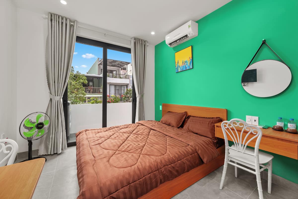 A bedroom with a large bed, a desk, a chair, a fan, and a view of the outside balcony.