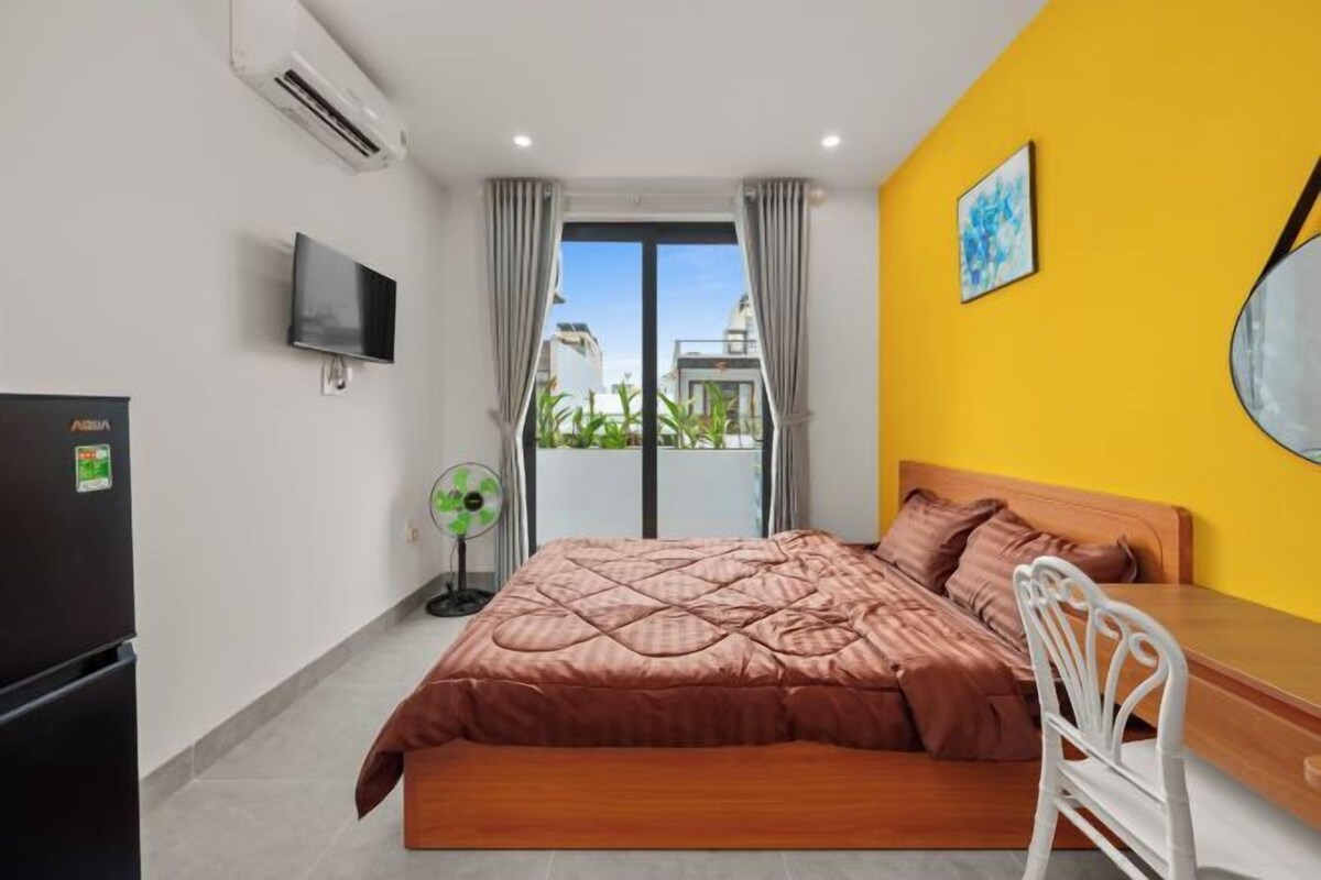 A bedroom with a bed, a TV, a fan, and a view of buildings outside.
