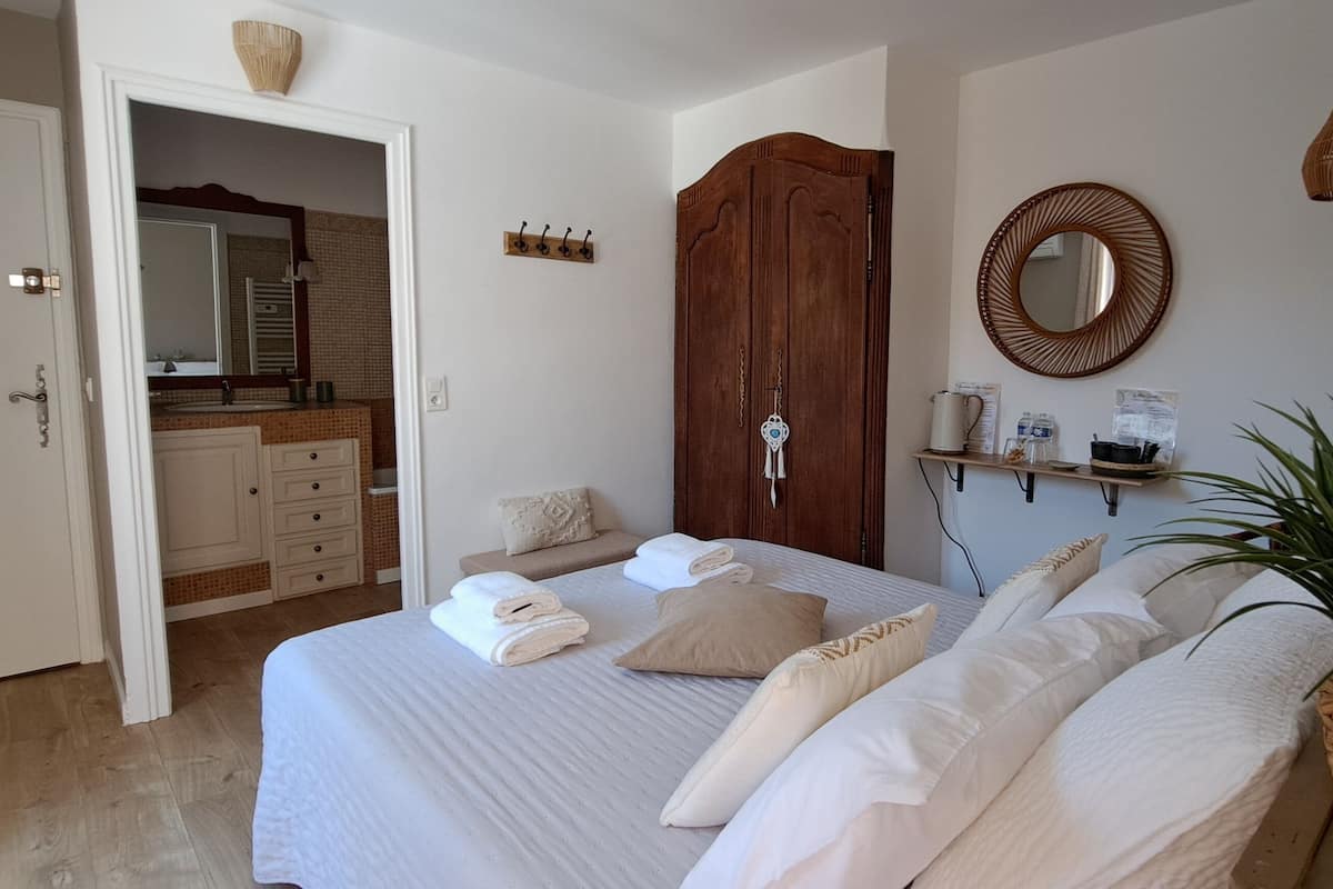 A bedroom with a bed, a wooden wardrobe, a mirror, a shelf with toiletries, and a potted plant.