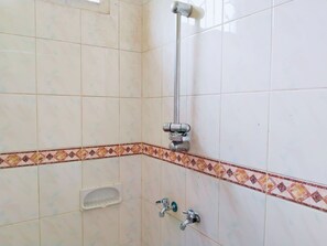 Baño con azulejos blancos, ducha, cabezal de ducha manual y un lavamanos pequeño con jabonera.