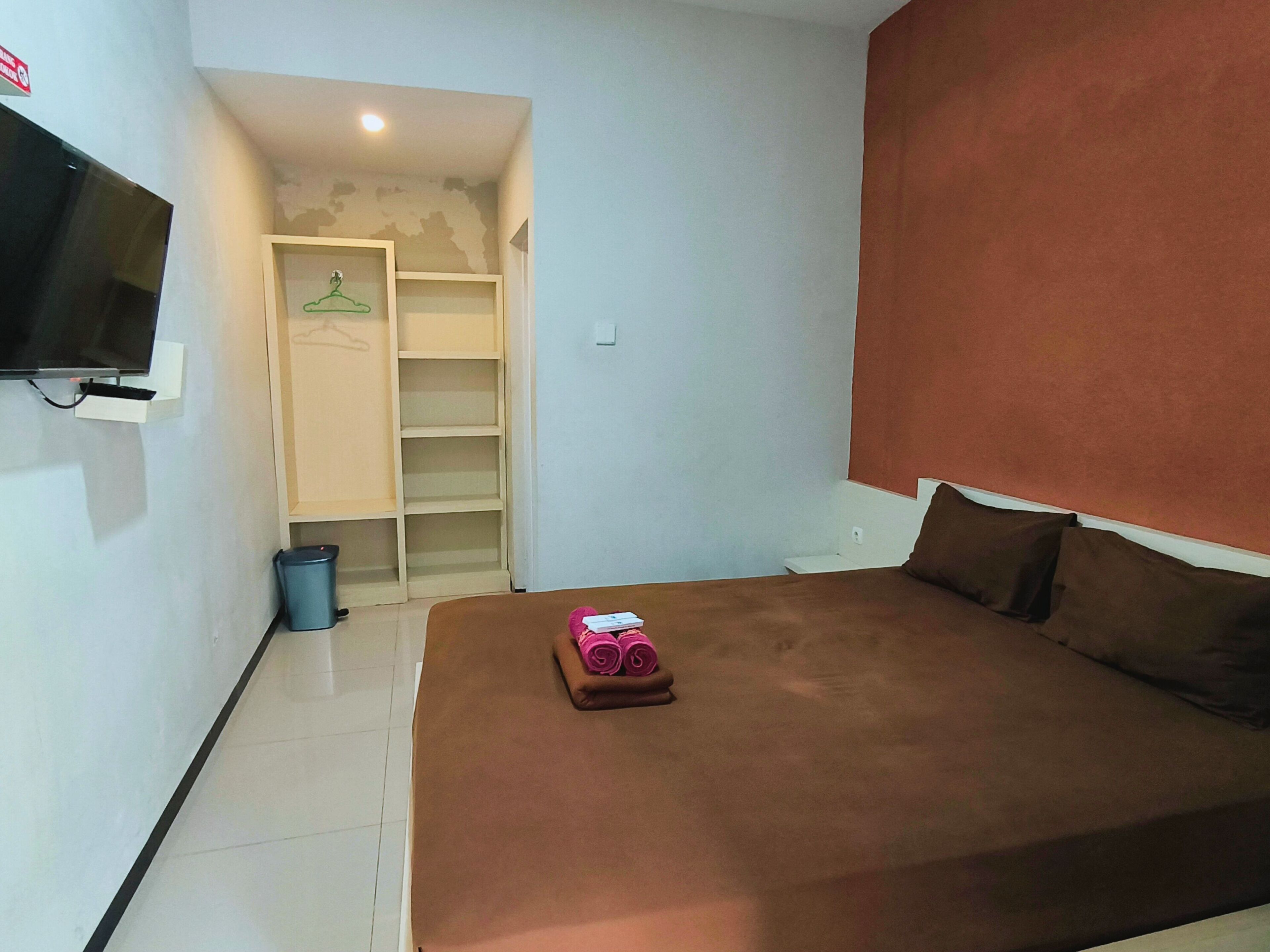 Un dormitorio con una cama, un televisor montado en la pared, un armario y un cubo de basura pequeño.