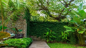 A wooden pathway leading through a lush garden with a stone wall and various tropical plants. - Peaceful Retreat with a Lush Garden & Open-Air Patio (Bantul)
