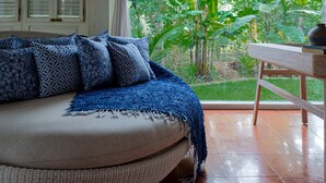 A room with a sofa, patterned cushions, a woven basket, a wooden table, and a view of lush greenery outside. - Peaceful Retreat with a Lush Garden & Open-Air Patio (Bantul)