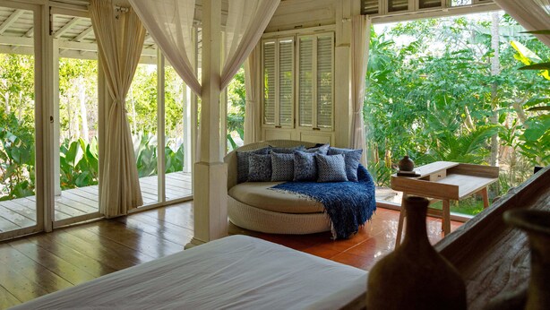 A room with a bed, a sofa with cushions, a small table, and a view of lush greenery outside. - Peaceful Retreat with a Lush Garden & Open-Air Patio (Bantul)