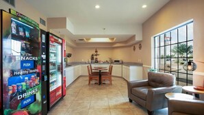 A hotel lobby with vending machines for snacks and drinks, a dining area with a round table and chairs, and a view through large windows.