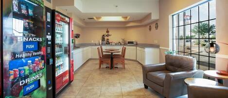 A hotel lobby with vending machines for snacks and drinks, a dining area with a round table and chairs, and a view through large windows.