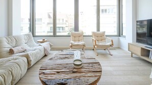 Un salon spacieux avec une grande fenêtre, une table basse ronde, un canapé, deux fauteuils et un téléviseur.