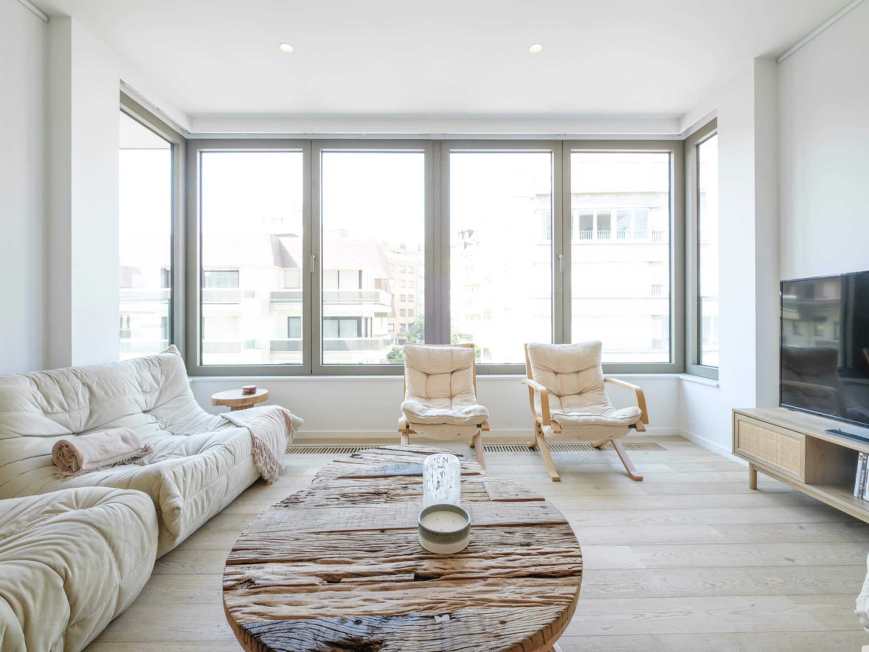 A spacious living room with a large window, a round coffee table, a sofa, two armchairs, and a television.