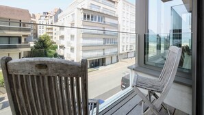 Un balcon avec une chaise en bois et une chaise blanche, donnant sur une rue bordée d’immeubles.
