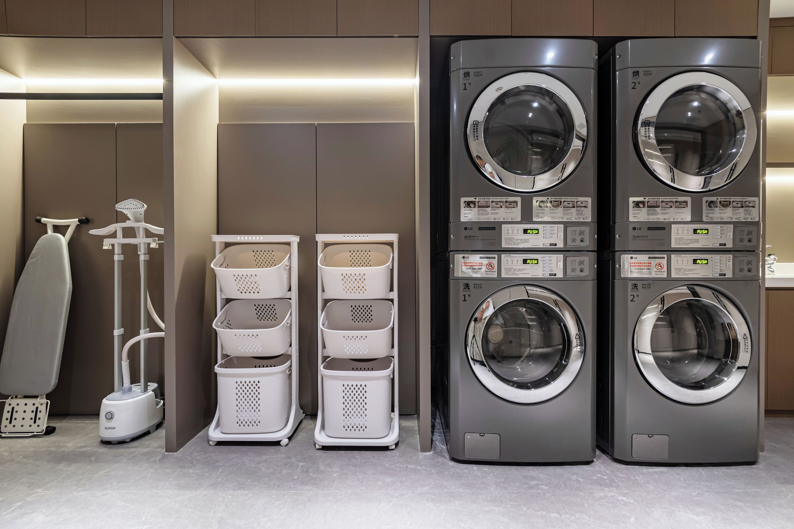 A laundry area with two stacked front-loading washing machines, a drying machine, and a stack of white laundry baskets.