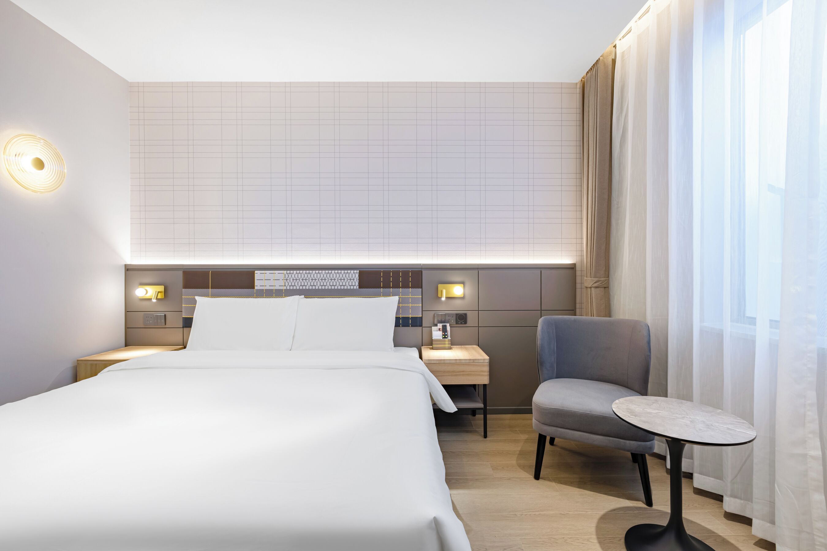 A modern hotel room with a large bed, a small table, a chair, and a nightstand with a lamp.
