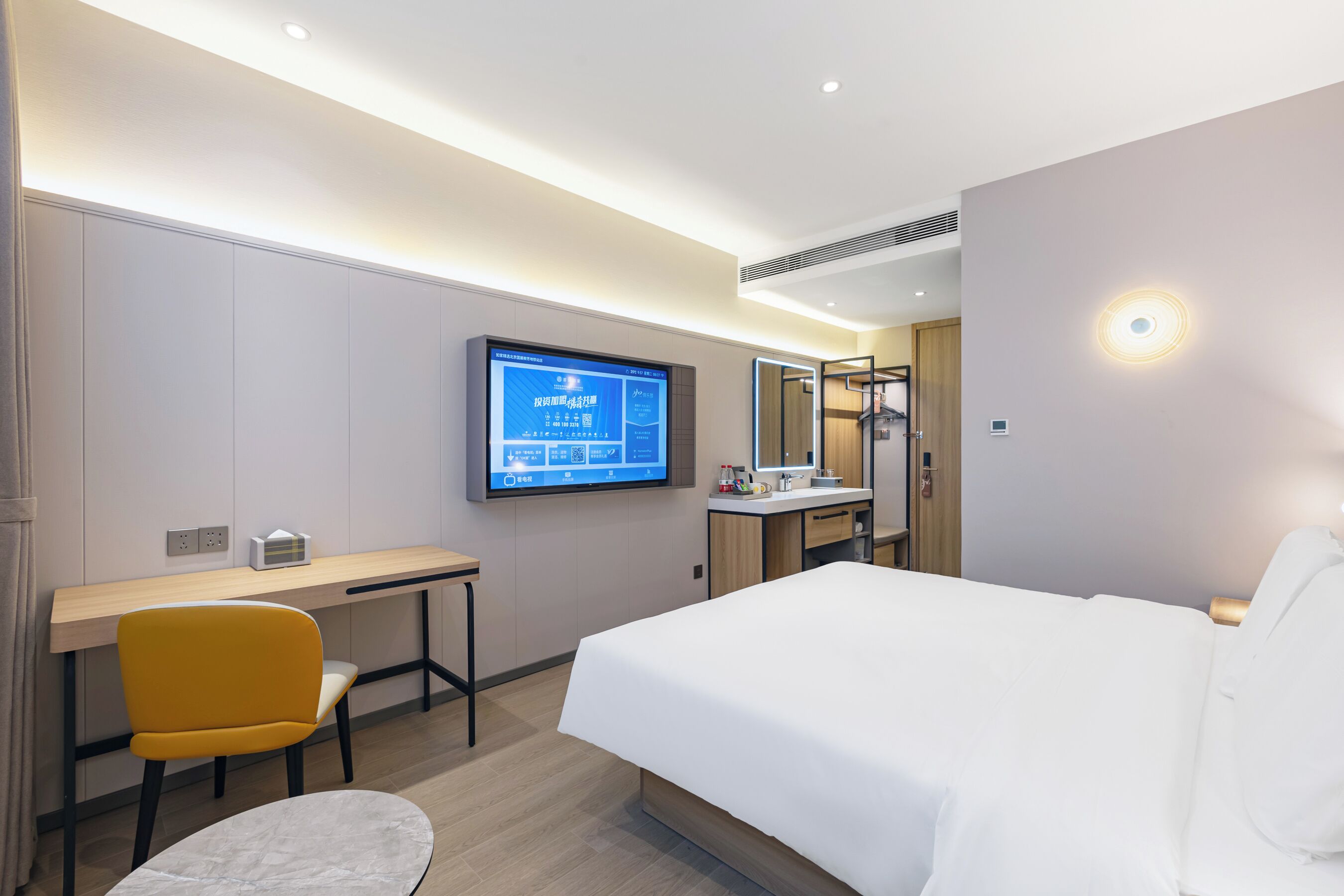 A modern hotel room with a bed, a desk, a chair, a TV mounted on the wall, and a bathroom with a mirror and sink.