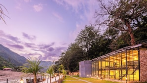 A modern building with large glass windows, a brick exterior, and a pathway leading to a seating area with tables and chairs. - DLS Divine River Resort and Spa (Rishikesh)