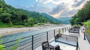 A riverside terrace with outdoor seating and a view of a river and mountains. - DLS Divine River Resort and Spa (Rishikesh)
