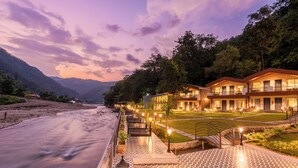 A hotel with a terrace overlooking a river, a well-lit garden, and a view of the surrounding hills. - DLS Divine River Resort and Spa (Rishikesh)