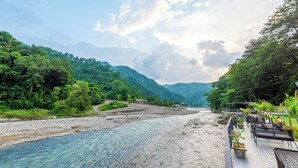 A riverside view with a paved walkway and seating area, surrounded by lush greenery and mountains. - DLS Divine River Resort and Spa (Rishikesh)