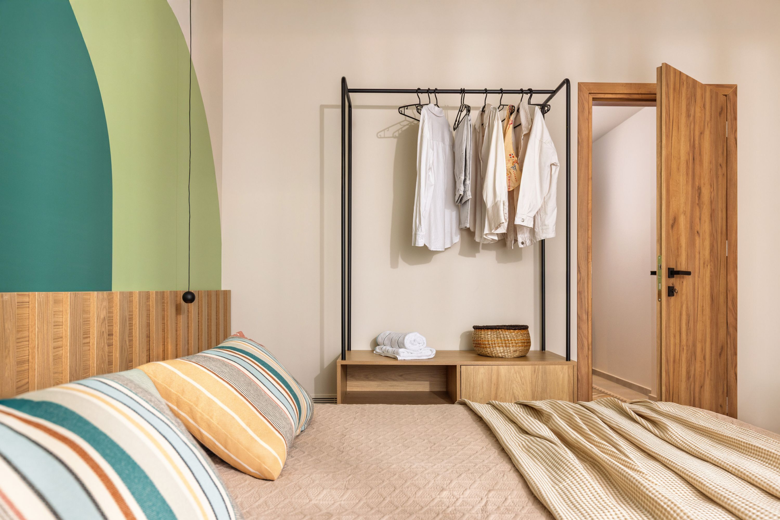 A bedroom with a bed, a hanging clothes rack, a wooden wardrobe, and a small wooden bench.