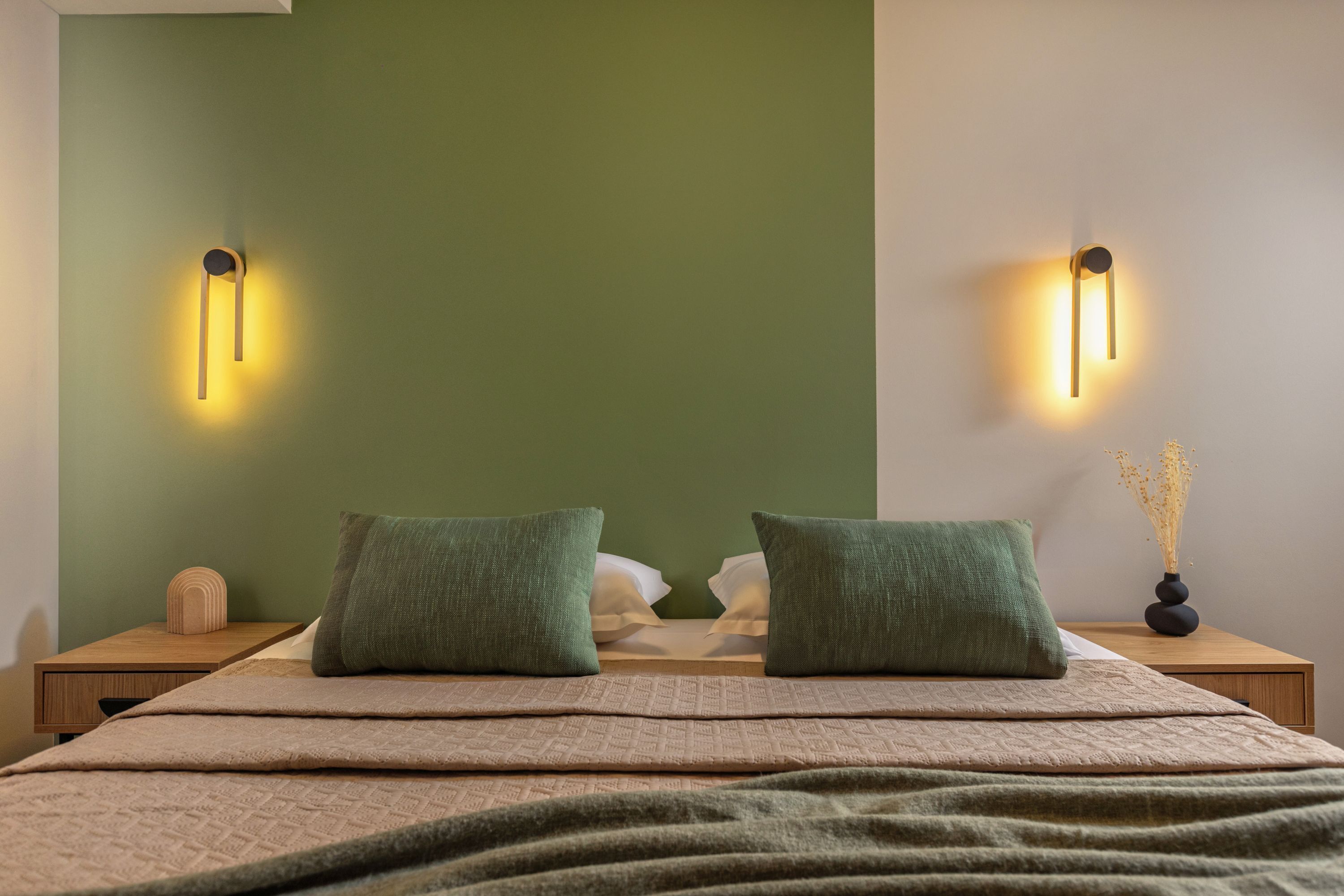 A neatly made bed with two pillows, a bedside table with a lamp, and a vase with dried flowers on the nightstand.