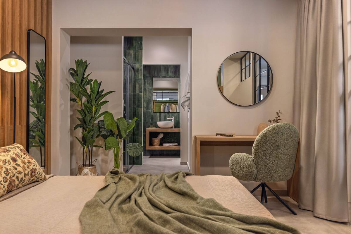 Une chambre moderne avec un grand lit, un miroir rond, un bureau et une chaise. On y trouve une plante et une étagère avec des serviettes et un bol.