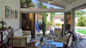 A sunlit patio with a sofa, chairs, a table, and a glass door leading to an outdoor area with a pool. - House in Provence (Chateauneuf de gadagne)